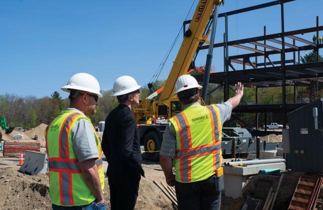 Two men stand with the U N E President as they survey the construction site for the upcoming health sciences building